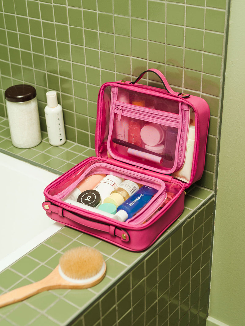 Water-resistant CALPAK Clear Cosmetics Case in pink Dragonfruit being used in a green-tiled bathroom; CCC2001-DRAGONFRUIT