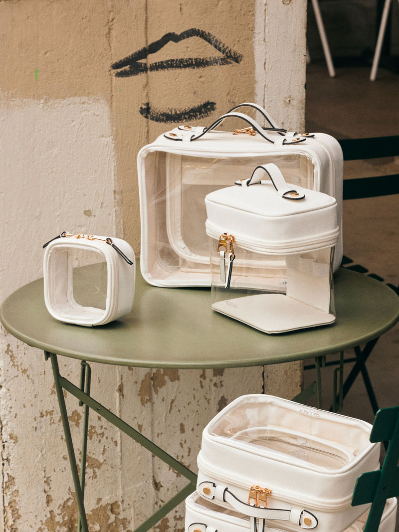 CALPAK Clear Train Case in White on a green table, with other CALPAK Clear Cosmetic Cases in white