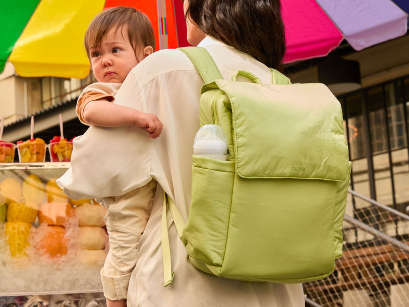 Mother holding her toddler in her arms while wearing the CALPAK Diaper Backpack in Lime in front of a fruit stand