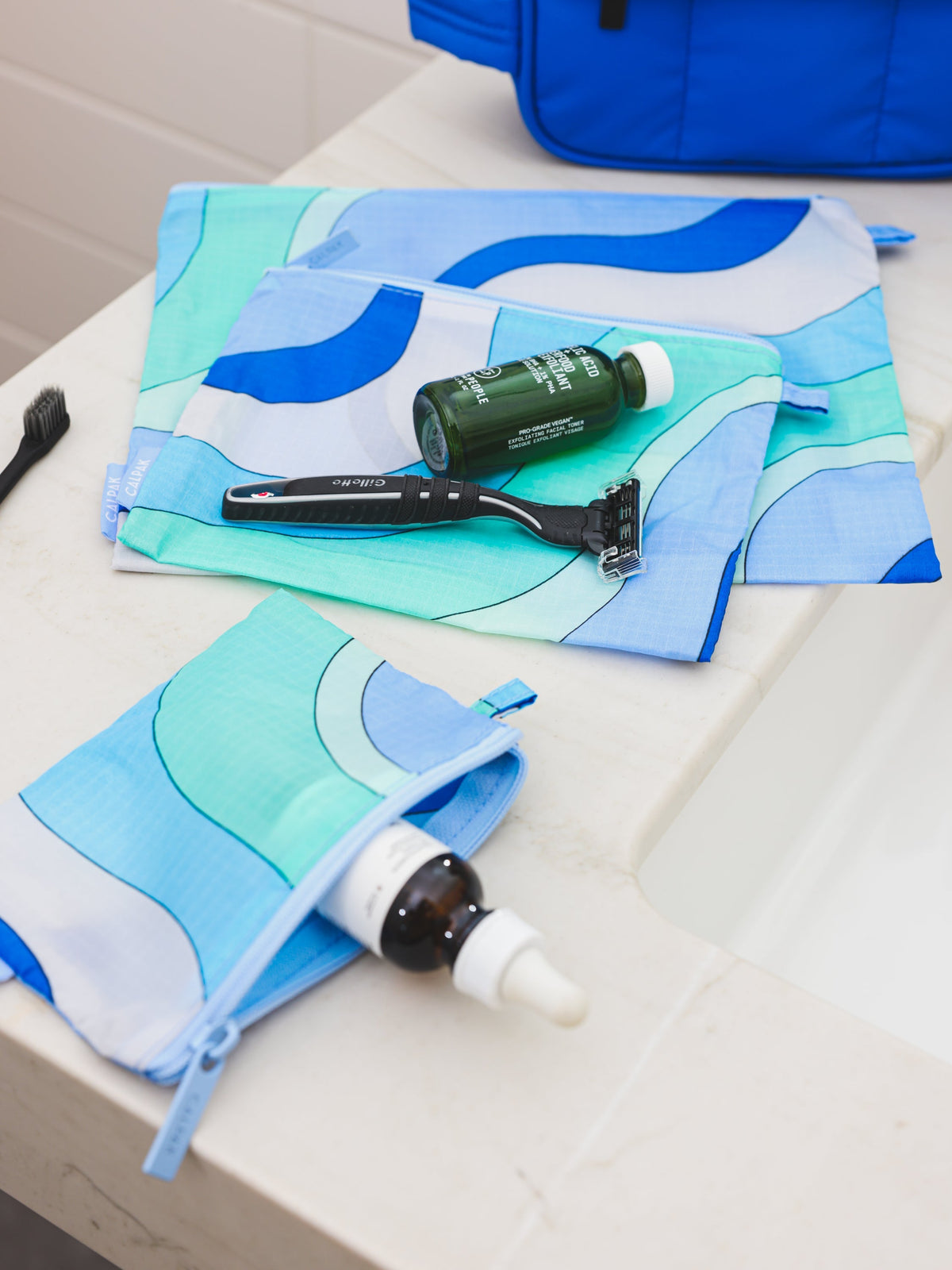 From CALPAK's Compakt collection, these 'Groovy Blue' zippered nylon pouches are displayed on a bathroom sink, offering stylish organization for your essentials; KZB2001-GROOVY-BLUE