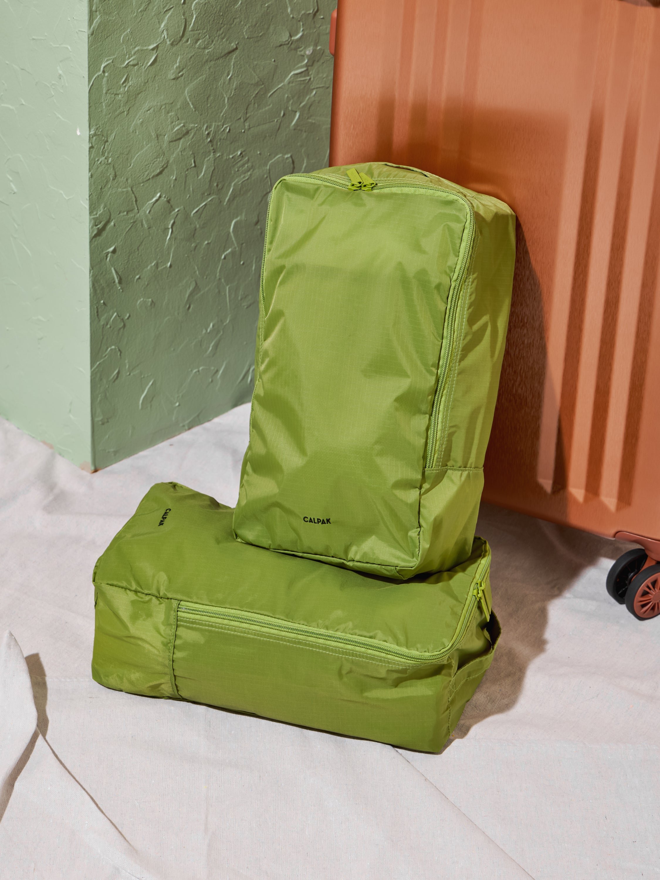 CALPAK Compakt Shoe Bag in green palm stacked on one another and leaning against CALPAK Ambeur Carry-On Luggage in copper
