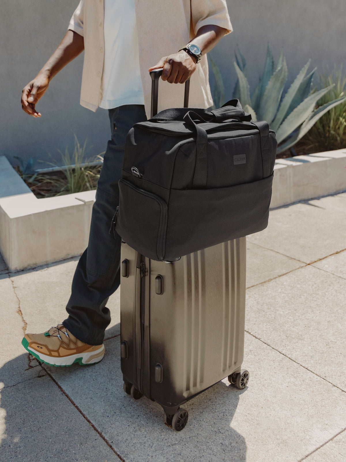 Model rolling CALPAK Ambeur Carry-On Luggage with CALPAK Connect Laptop Duffel fastened at luggage trolley sleeve in black; DCN2401-BLACK