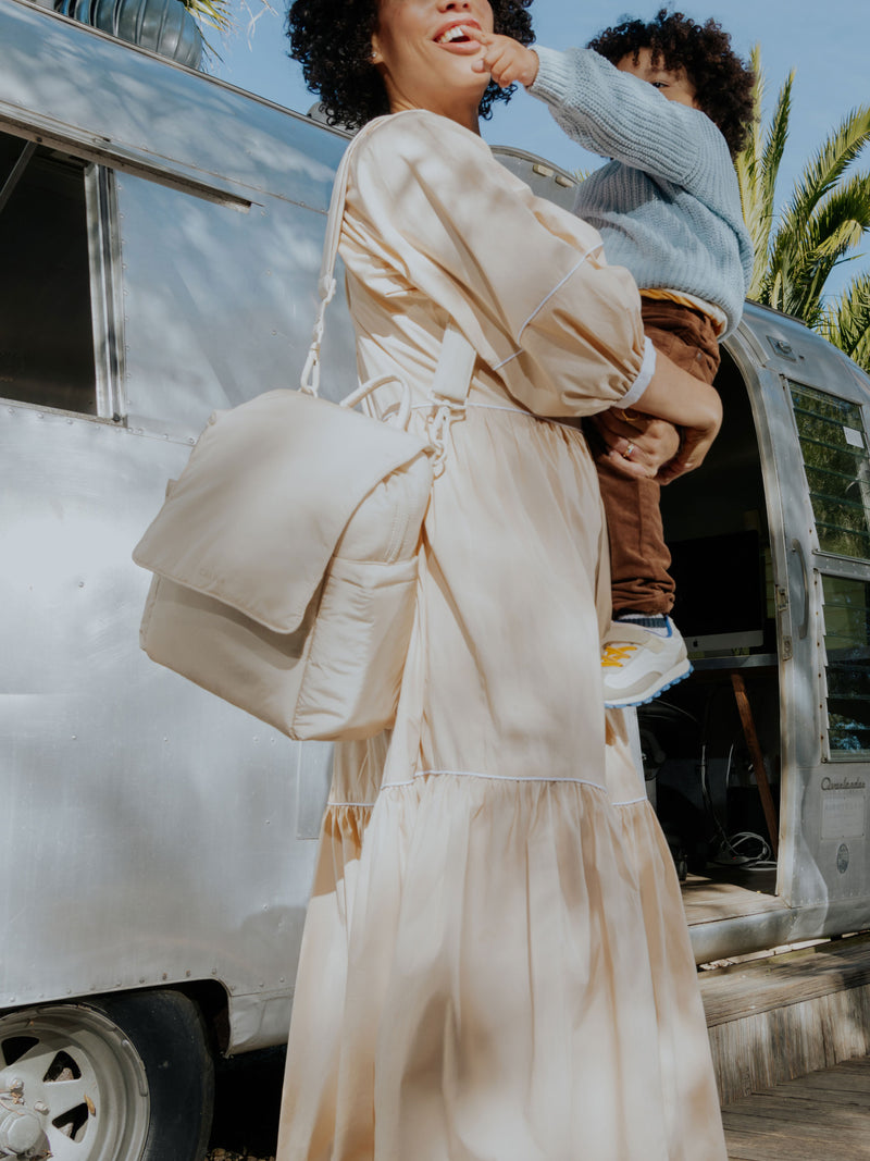 The Convertible Mini Diaper Backpack in oatmeal, shown in a lifestyle setting worn on the shoulder of a woman carrying a young child