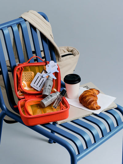 CALPAK x Dedcool Collaboration of the Small Clear Cosmetic Case in orange Papaya with toiletry items and perfumes  on a airport chair; CCM2001-DEDCOOL view 2