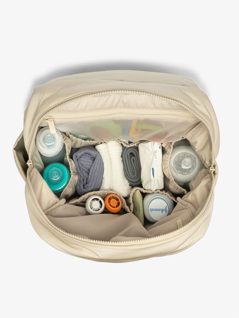 An open diaper backpack in an oatmeal color is displayed, showing the interior layout. The bag features a spacious main compartment with several pockets for organization, including a padded laptop sleeve. The inside lining is light-colored, making it easy to see and access items.