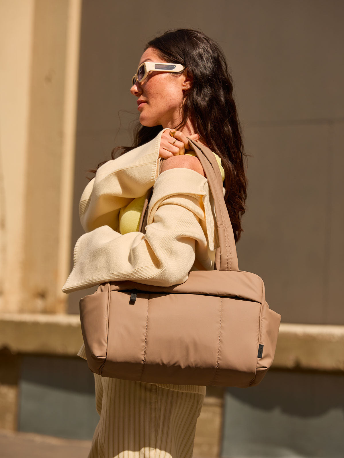 CALPAK Mini Duffel in brown Chocolate  with puffy exterior, interior and exterior pockets, and padded shoulder straps being worn by a model; DLM2501-CHOCOLATE