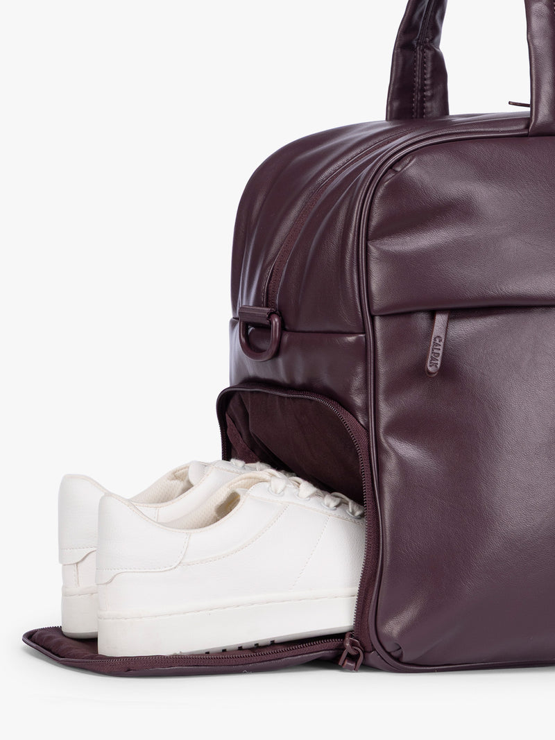 Side shoe compartment of a dark burgundy CALPAK Luka Duffel in vegan leather with dual top handles and a front zippered pocket, photographed against a clean white background
