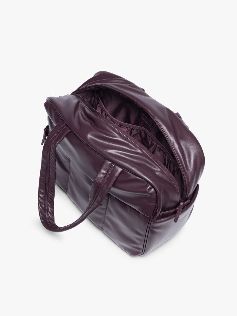 Side view with zipper open showing inside of A dark burgundy CALPAK Luka Duffel in vegan leather with dual top handles and a front zippered pocket, photographed against a clean white background
