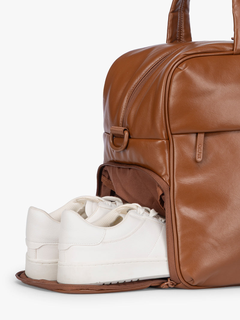 Side shoe compartment of Honey Brown CALPAK Luka Duffel in vegan leather with dual top handles and a front zippered pocket, photographed against a clean white background