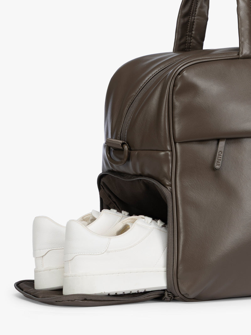 Side shoe compartment of A dark brown Mushroom CALPAK Luka Duffel in vegan leather with dual top handles and a front zippered pocket, photographed against a clean white background