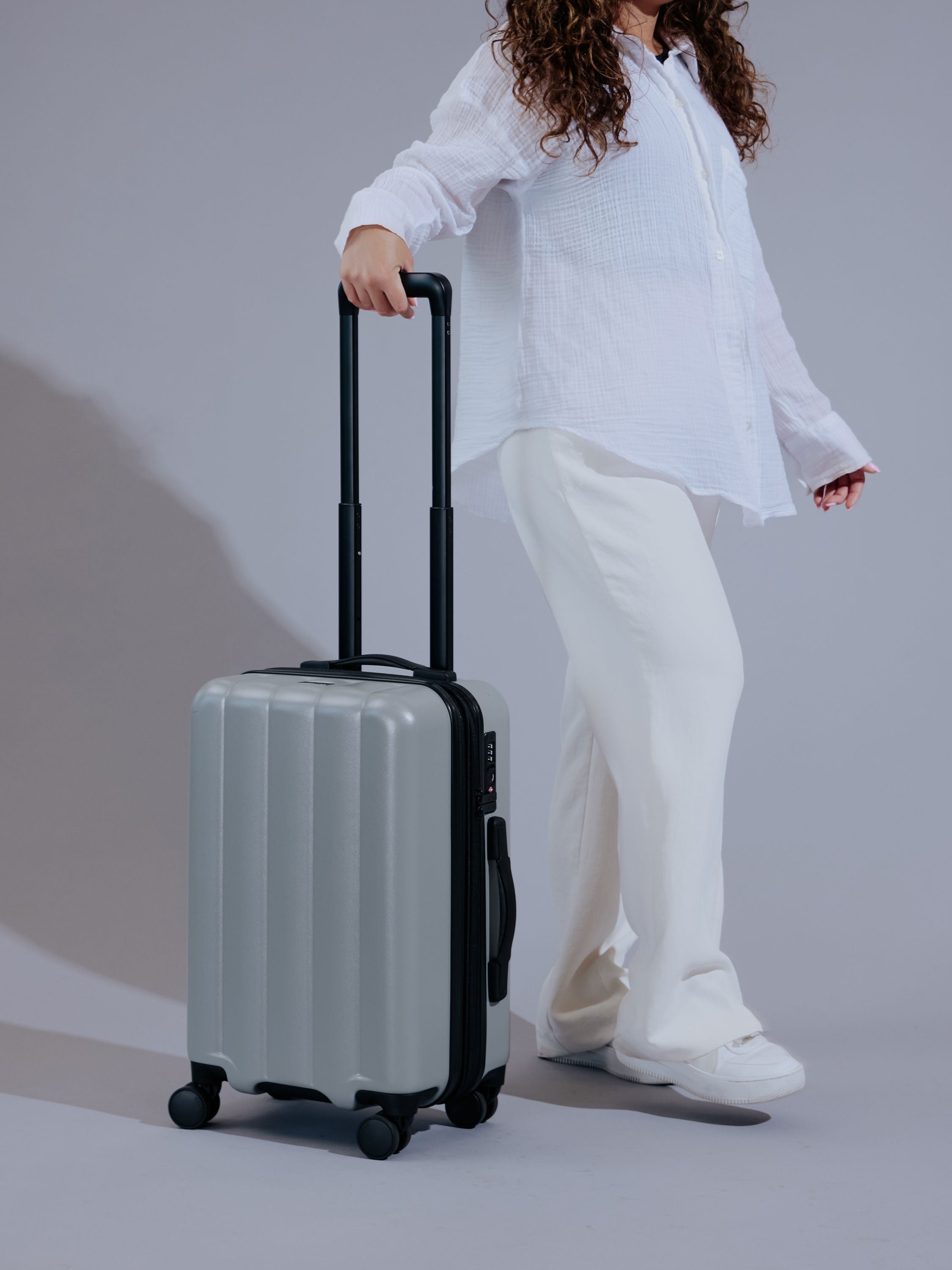 Model rolling smoke gray CALPAK Evry Carry-On Luggage and displaying dual spinner wheels