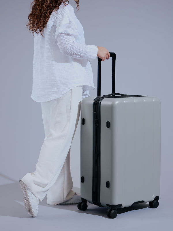 Model standing besides durable CALPAK Large Luggage in smoke featuring dual spinner wheels