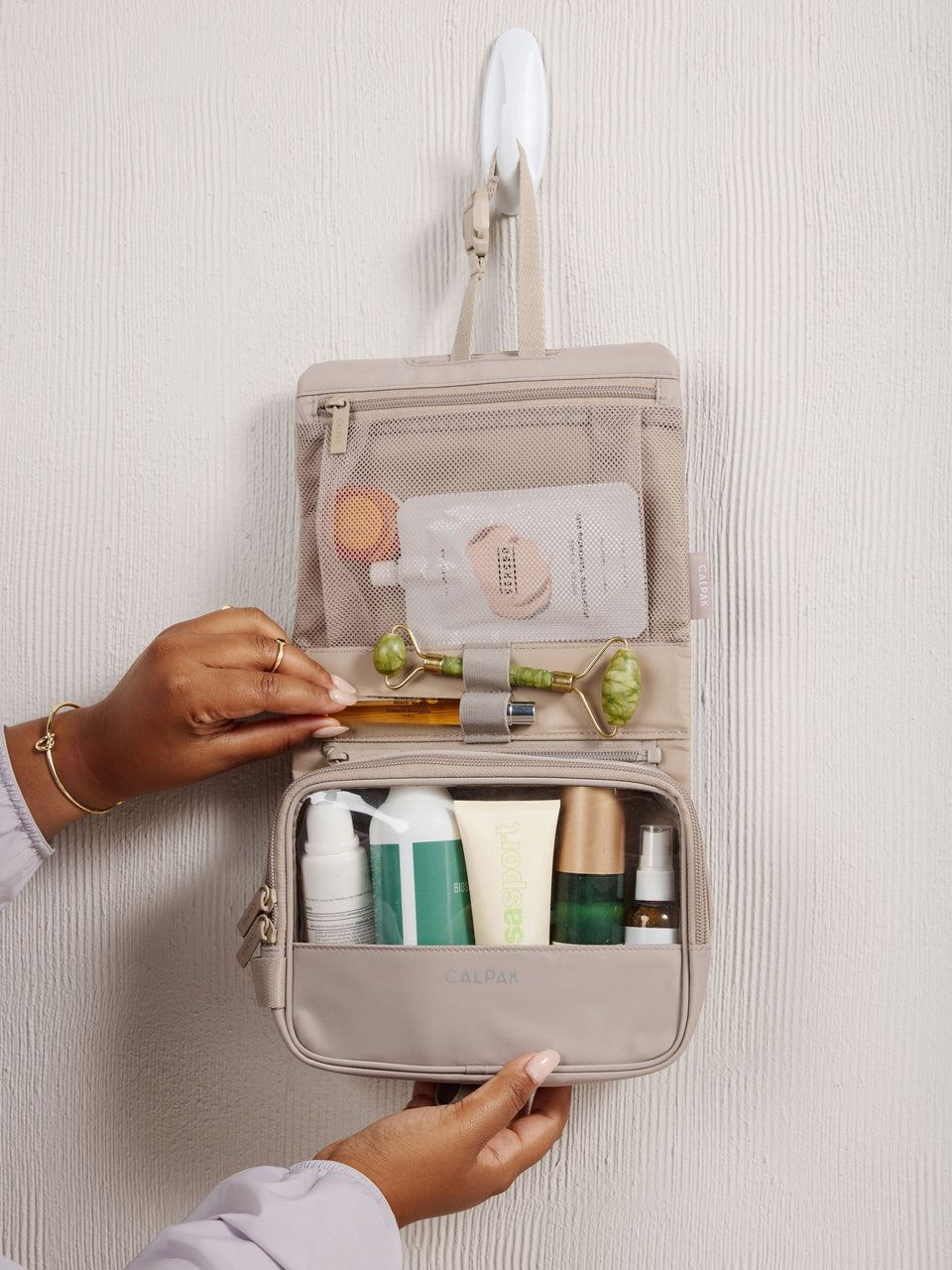 Model placing perfume bottle within elastic loops of CALPAK Fit gym toiletry bag full of toiletries in grayish beige; TFH2401-GREIGE, TFH2401-GREIGE-GRIN, BTBF2401-GREIGE