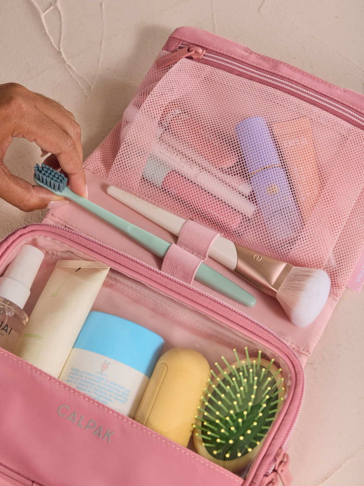 Model removing toothbrush from elastic loop of CALPAK Fit Hanging Dopp Kit in tea rose with toiletries packed within gym toiletry bag; TFH2401-TEA-ROSE, BTBF2401-TEA-ROSE