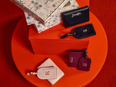 A styled flat-lay of CALPAK Luggage Tag and Passport Holder Set. Showing gift boxes arranged on a circular red platform against a textured red background. The set includes a cream toile-print box, black tags with gold monogram initials, and two burgundy tags with purple monogram initials