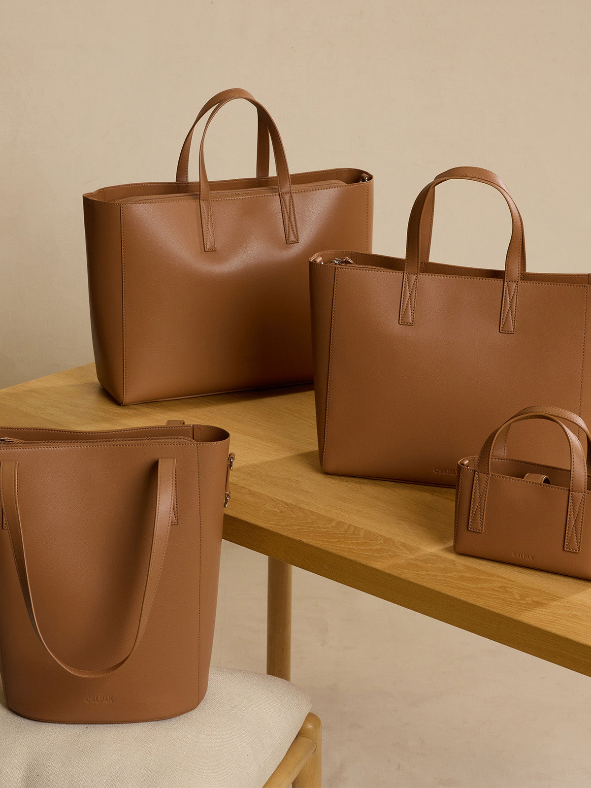 Four tan CALPAK Haven tote bags in different sizes arranged on a light wood table against a warm beige wall.; ATS2501-TOFFEE