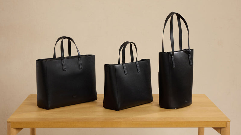 Three black CALPAK Haven totes of different sizes on a wooden table with a beige background