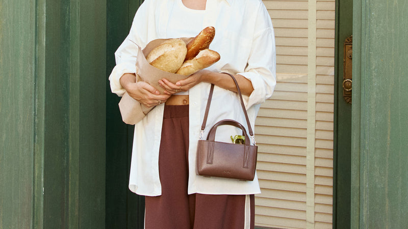 Person holding bread and CALPAK Mini Crossbody Haven Bag in dark brown Espresso\