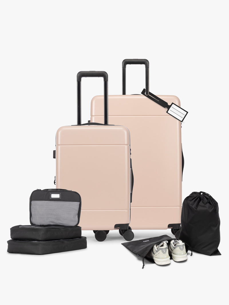 Opened CALPAK Hue Starter Bundle in light pink Rose Quartz that comes with Large and Carry On Luggage, Luggage Tag, Two Black Pouches, and Three Black Packing Cubes shown on a white background