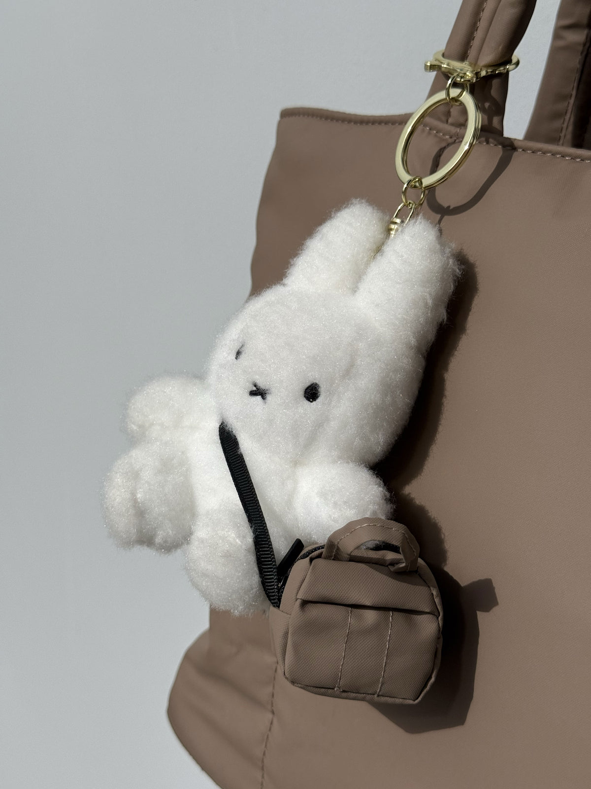 White fluffy bunny charm attached to a bag with the CALPAK Teeny Tiny Luka Duffel with strap in brown Chocolate;TTD2501-CHOCOLATE