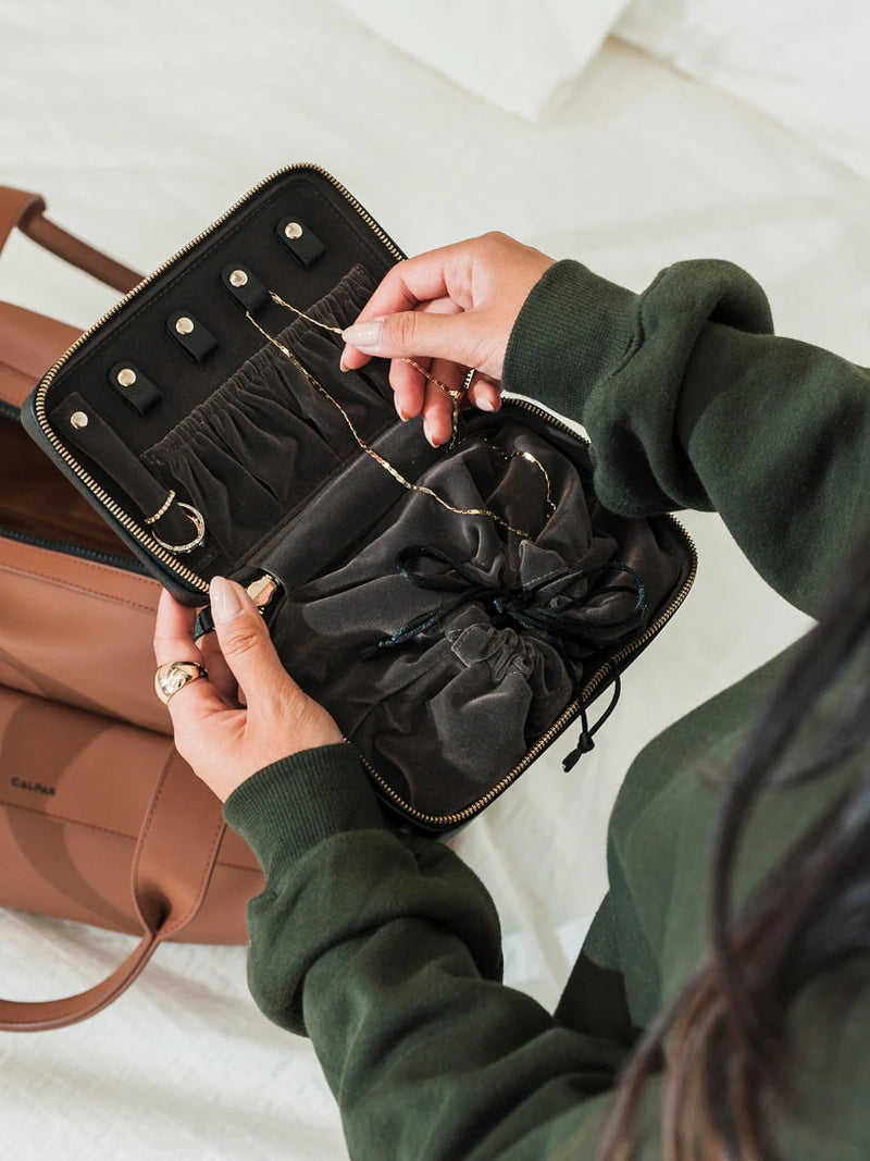 A top view of the CALPAK black jewelry case with a suede interior, pouch, and ring holders. A necklace is placed inside, using the snap tab closure.