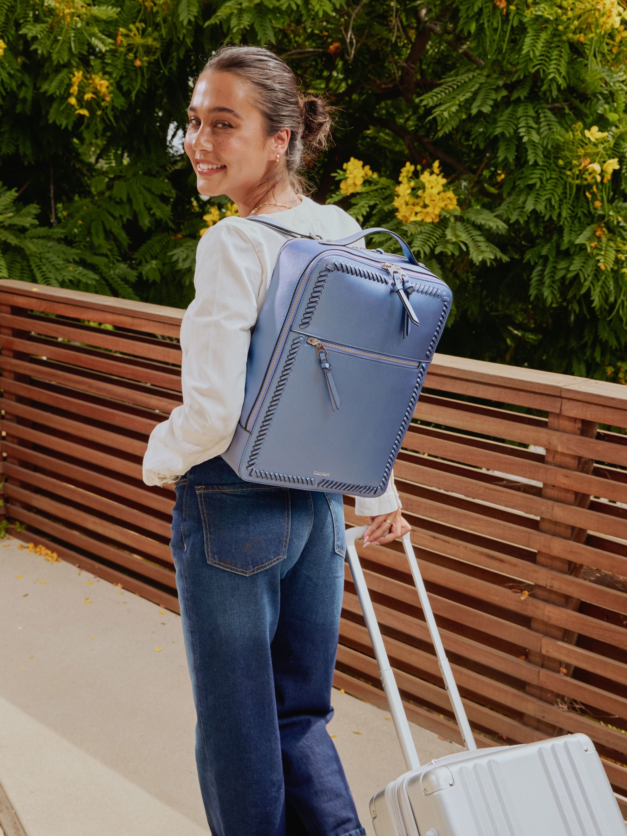 Model wearing CALPAK Kaya Laptop in metallic blue stargaze on back and rolling Ambeur Carry-On Luggage in silver
