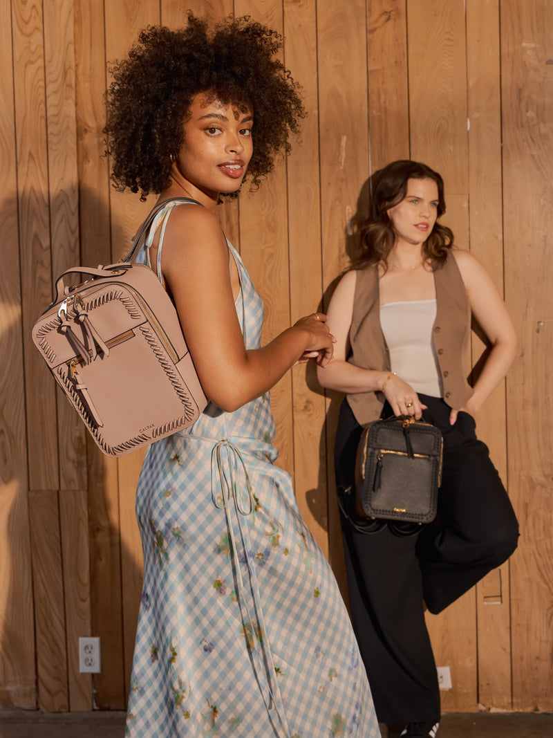  A woman is carrying a Kaya mauve mini backpack over one shoulder while another woman in the background carries the Kaya mini backpack in black in the hand. The backpack has a structured, compact design with a front pocket, gold-tone zipper accents, and a top handle.