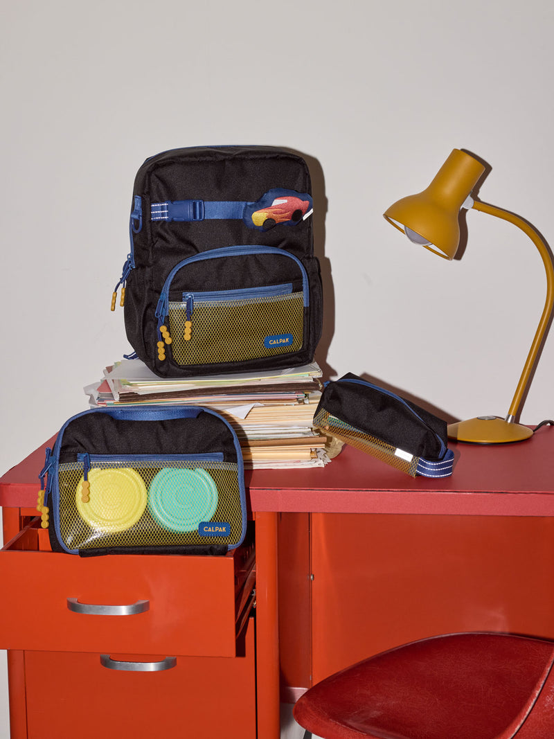 CALPAK Kids Small Backpack in Black for ages 3-5 is built for your little one’s big days with blue zippers and yellow mesh front pocket, multiple interior pockets for an 11” tablet or laptop, and padded comfort straps on a red desk