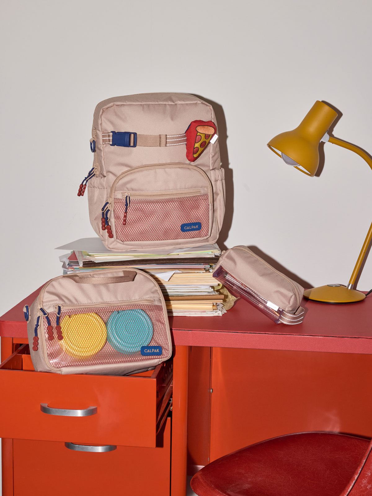 CALPAK Kids Small Backpack in brown and tan Khaki for ages 3-5 is built for your little one’s big days with dark brown and tan zippers and red mesh front pocket, multiple interior pockets for an 11” tablet or laptop, and padded comfort straps on a red desk; CKBS2501-KHAKI