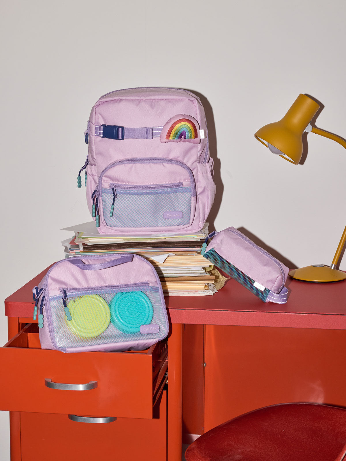 CALPAK Kids Small Backpack in lavender pink Orchid Pink for ages 3-5 is built for your little one’s big days with lavender pink  zippers and blue mesh front pocket, multiple interior pockets for an 11” tablet or laptop, and padded comfort straps on a red desk; CKBS2501-ORCHID-PINK