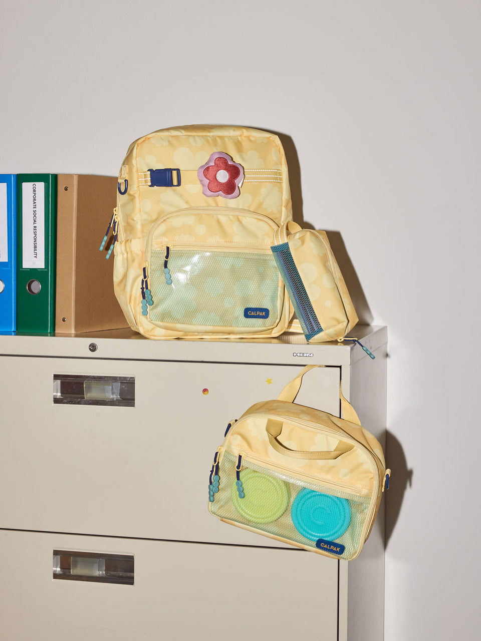 CALPAK Kids Medium Backpack in yellow with tan flower print in Yellow Bloom for ages 5-8 is built for your little one’s big days with yellow zippers and blue mesh front pocket, multiple interior pockets, and padded comfort straps on a beige cabinet; CKBM2501-YELLOW-BLOOM