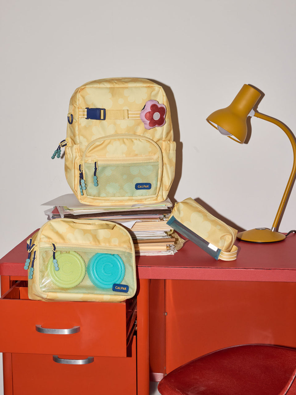CALPAK Kids Small Backpack in yellow with tan flower print in Yellow Bloom for ages 3-5 is built for your little one’s big days with yellow zippers and blue mesh front pocket, multiple interior pockets for an 11” tablet or laptop, and padded comfort straps on red desk; CKBS2501-YELLOW-BLOOM