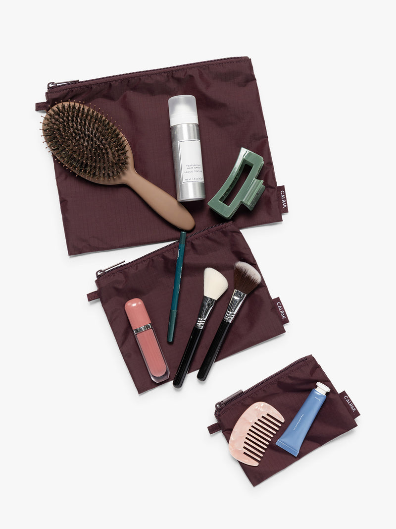 Items on top of the three burgundy Dark Cherry CALPAK Compakt Zippered pouches of different sizes, with items on top showing what fits inside, on a clean white background