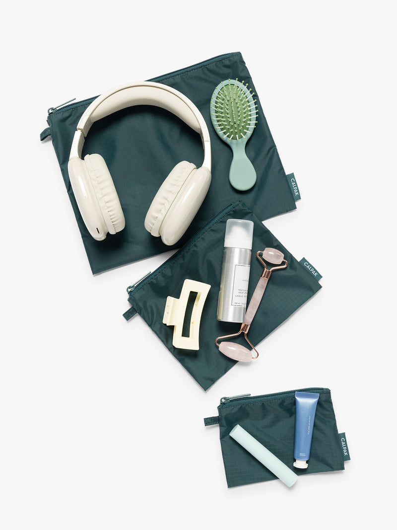 Items on top of the three dark green teal Kale CALPAK Compakt Zippered pouches of different sizes, with items on top showing what fits inside, on a clean white background