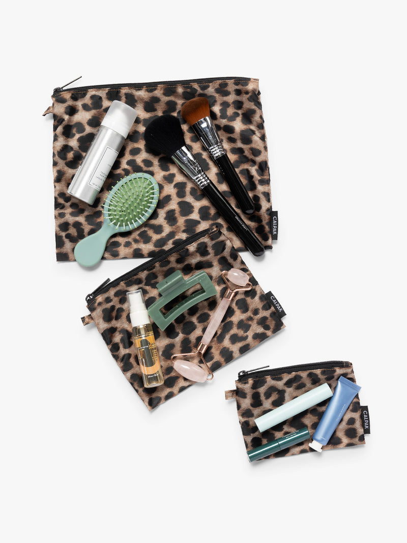 Items on top of the three tan and black animal print Leopard CALPAK Compakt Zippered pouches of different sizes, with items on top showing what fits inside, on a clean white background