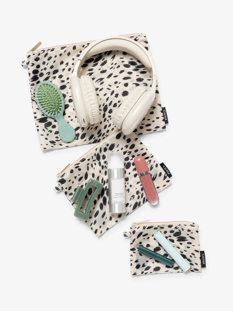 Items on top of the three white and black animal print Snow Leopard CALPAK Compakt Zippered pouches of different sizes, with items on top showing what fits inside, on a clean white background