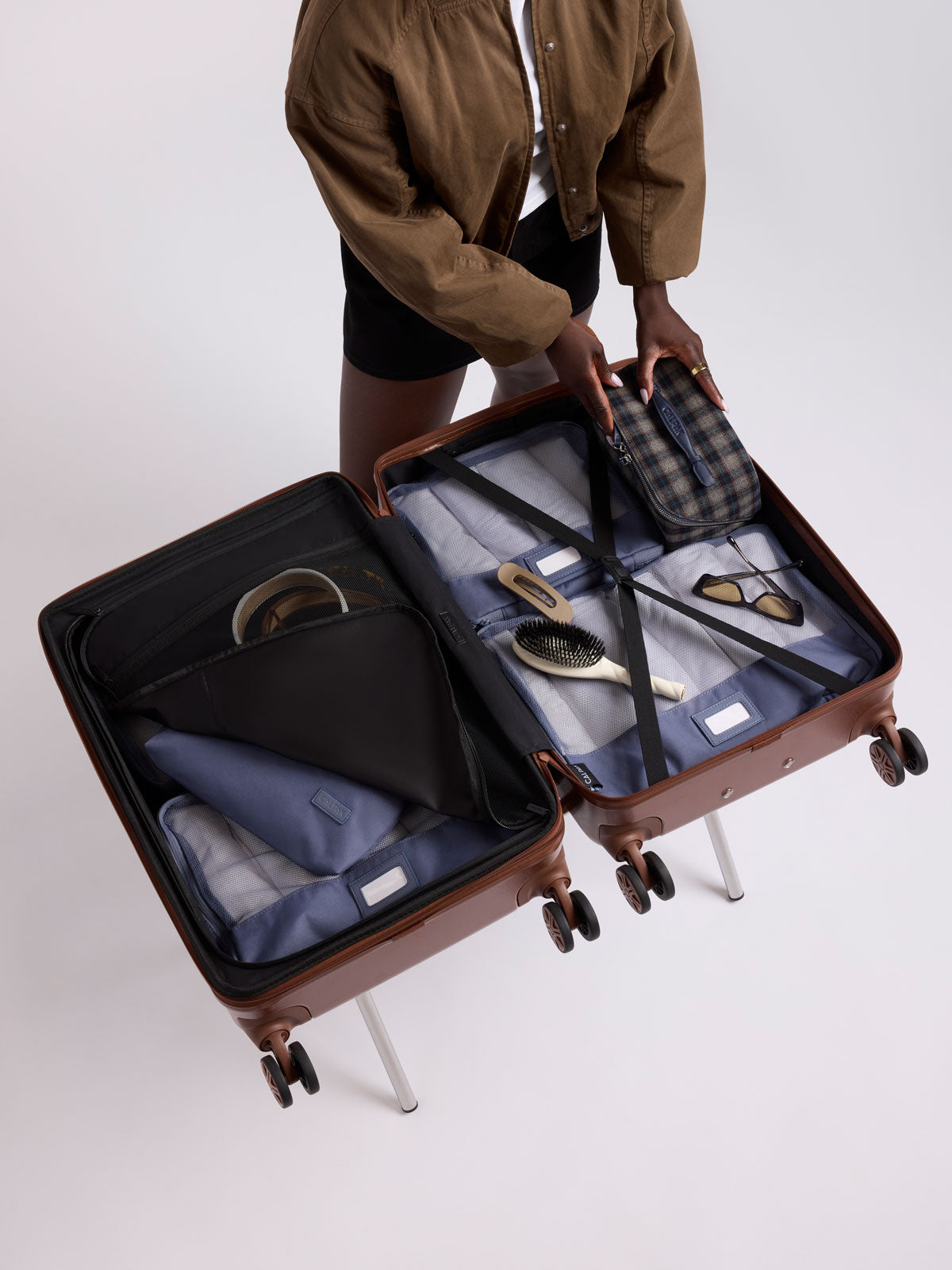 Model packing items into CALPAK Honey Brown Ambeur Carry On  hard-shell suitcase with a telescoping handle extended, shown from the front against a plain white background; LAM1020-HONEY-BROWN