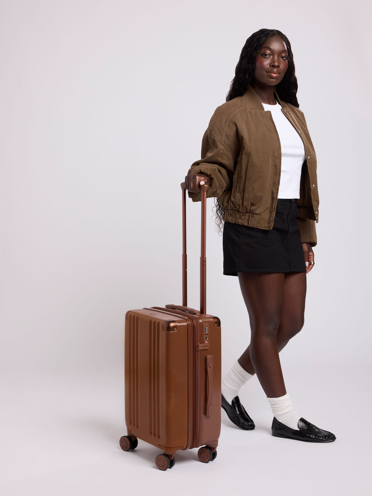 A woman stands in a studio holding the handle of CALPAK Honey Brown Ambeur Carry On  hard-shell suitcase with a telescoping handle extended while wearing a brown jacket, white top, black skirt, white socks, and black loafers against a light gray background; LAM1020-HONEY-BROWN