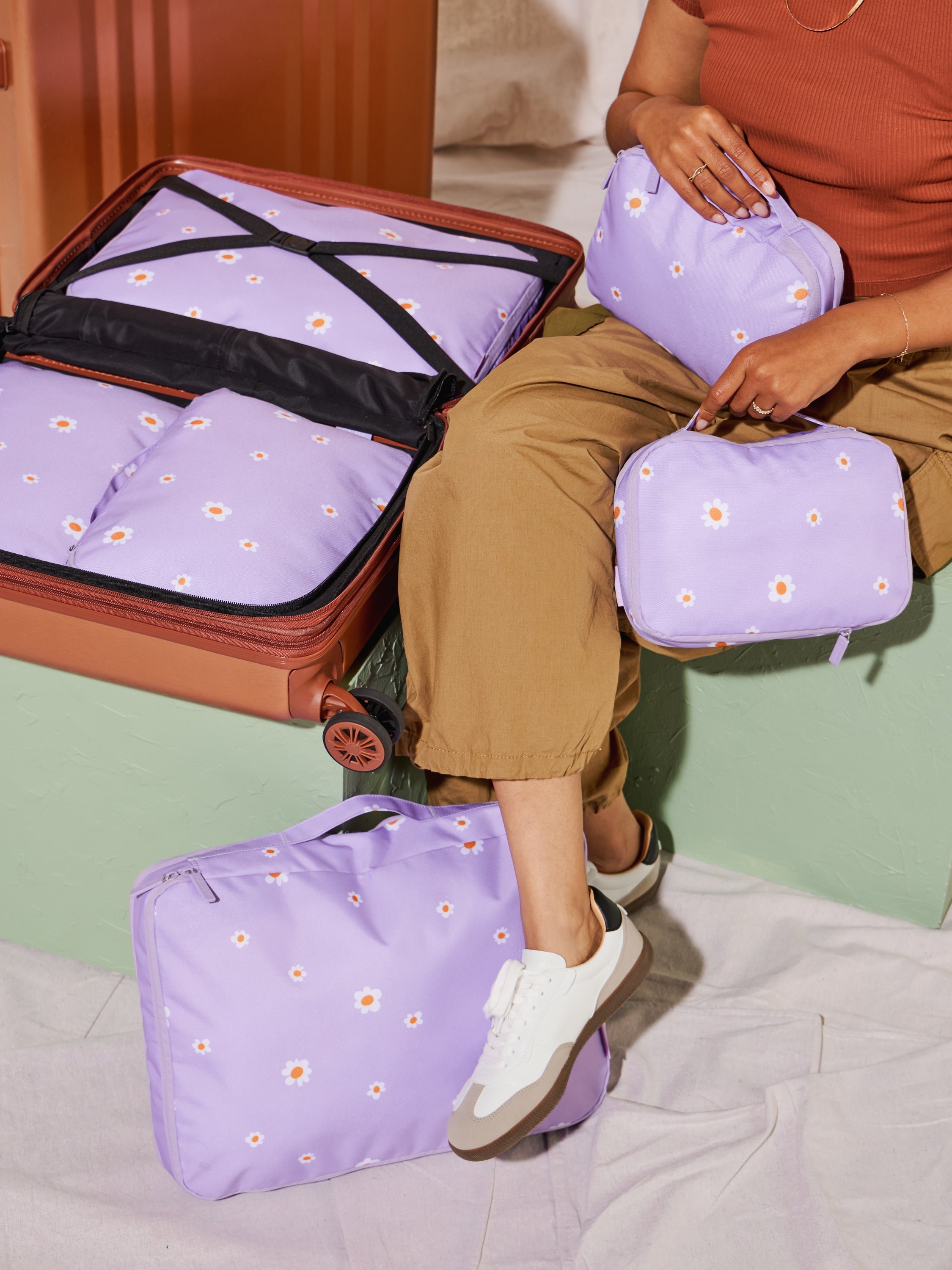 Model sitting besides suitcase packed with CALPAK Large Compression Packing Cubes and Medium Compression Packing Cubes while holding Small Compression Packing Cubes in orchid fields
