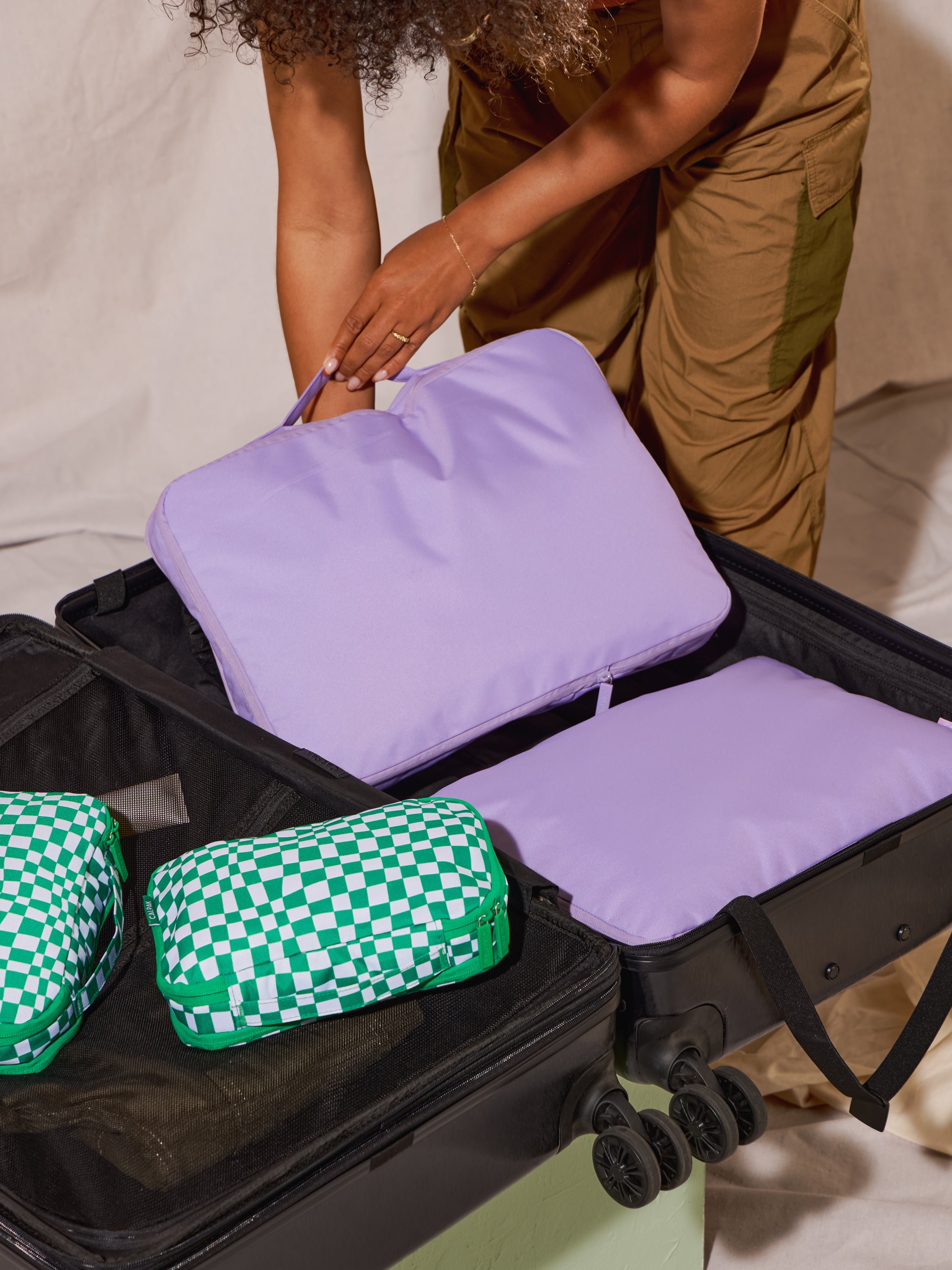 Model placing CALPAK Large Compression Cubes in orchid and CALPAK Small Compression Packing Cubes in green checkerboard within luggage