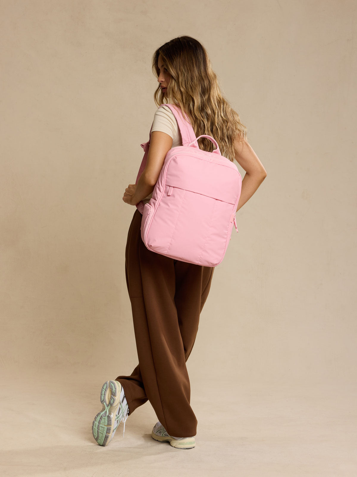 Woman carrying CALPAK Luka 15in Backpack in pink Blossom against a beige background; BPL2001-BLOSSOM