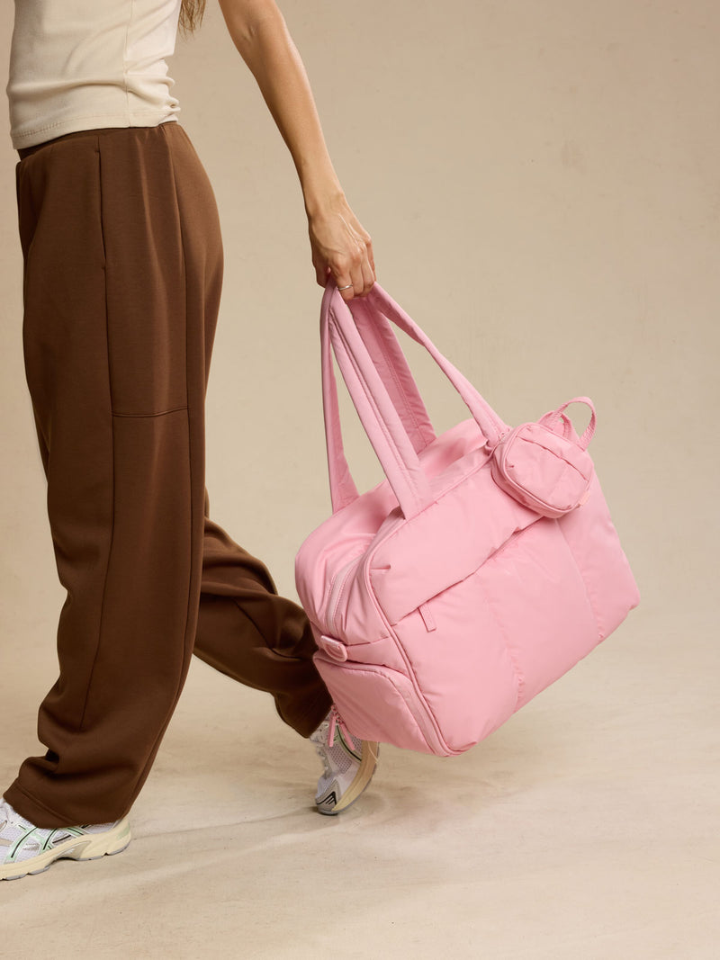 Woman carrying CALPAK Luka Duffel in pink Blossom against a beige background