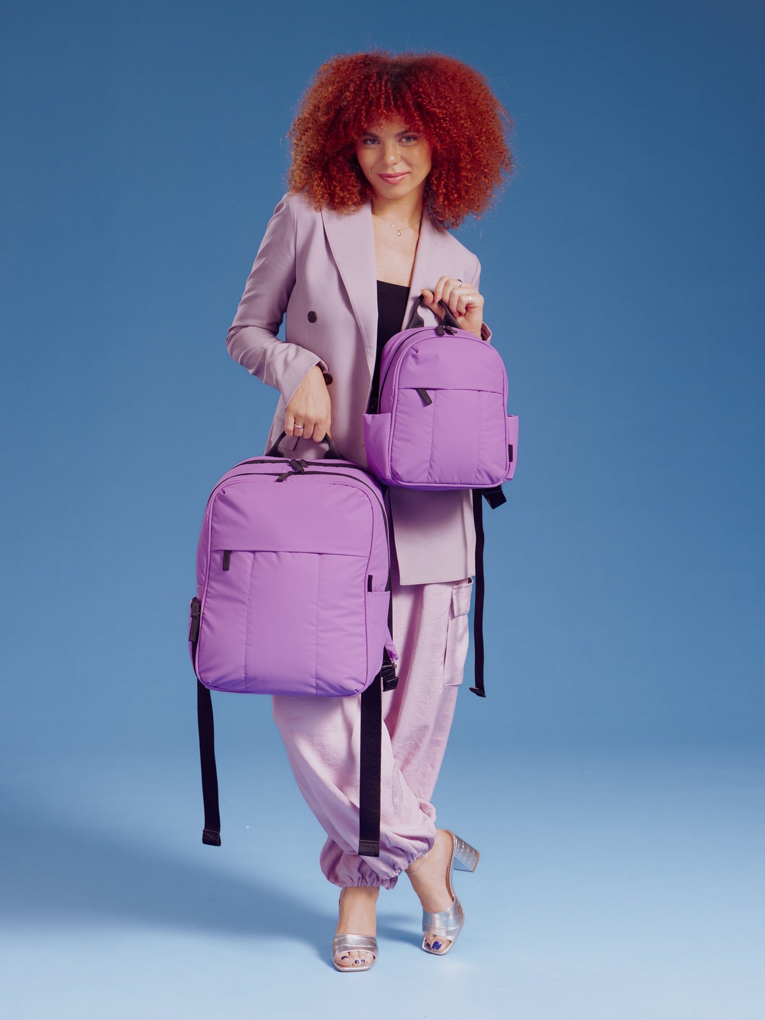 Model holding CALPAK Luka Mini Backpack and CALPAK 15 inch Laptop Backpack for size comparison in lavender