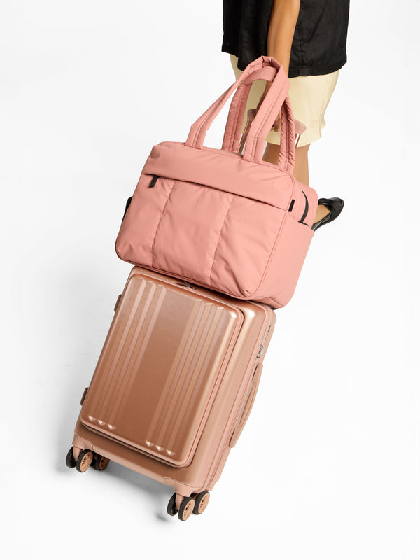 Model rolling Ambeur Front Pocket Carry-On Luggage in rose gold with peony Luka Duffel on top secured by trolley sleeve