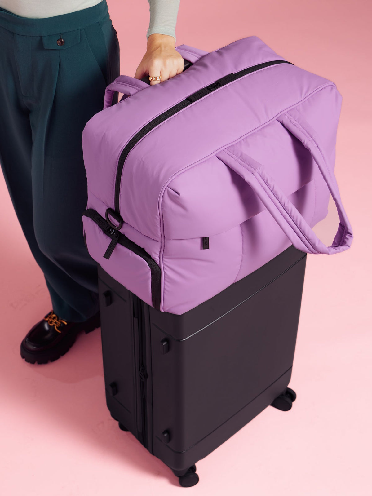 Model resting CALPAK Luka Large Duffel in lilac on top of CALPAK Hue Medium Carry-On Luggage in black; DLL2201-LILAC