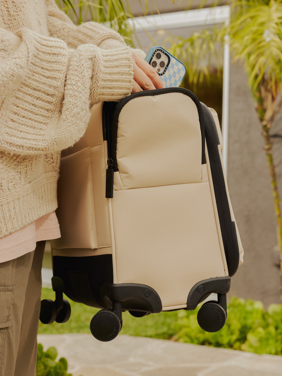 Model carrying CALPAK carry on Luka soft sided luggage with 360 spinner wheels and side pocket in ivory; LSM1014-OATMEAL