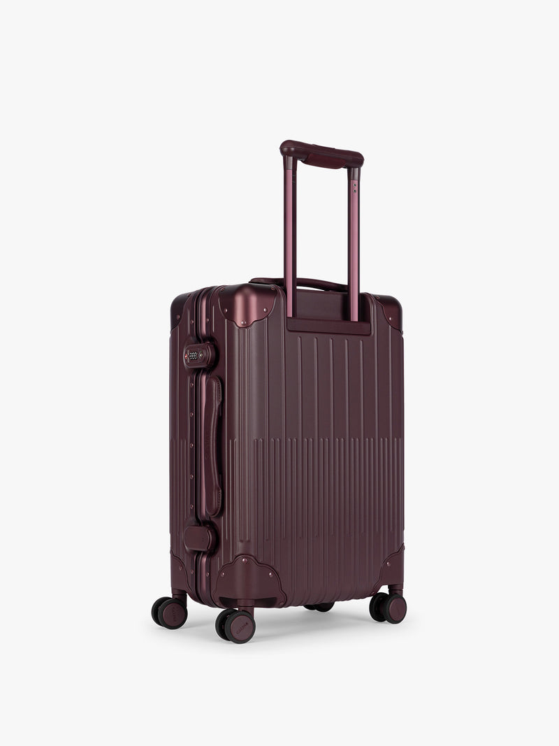 Backside of CALPAK Vault deep red Dark Cherry hard-shell spinner Carry On Luggage with a telescoping handle extended, featuring a deep red leatherette side handle and trim, set against a plain light gray background
