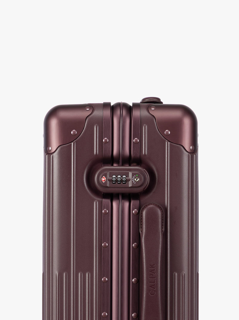 Closeup of the TSA-approved lock of CALPAK Vault deep red Dark Cherry hard-shell spinner Carry On Luggage with a telescoping handle extended, featuring a deep red leatherette side handle and trim, set against a plain light gray background