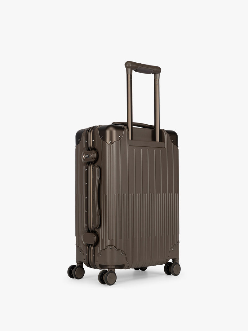 Backside of CALPAK Vault brown Mushroom hard-shell spinner Carry On Luggage with a telescoping handle extended, featuring a black leatherette side handle and trim, set against a plain light gray background