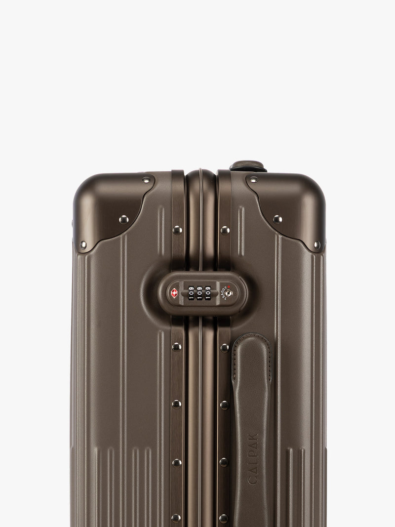 Close up of the TSA-approved lock of CALPAK Vault brown Mushroom hard-shell spinner Carry On Luggage with a telescoping handle extended, featuring a black leatherette side handle and trim, set against a plain light gray background